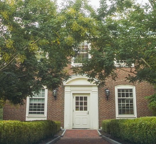 A tree and hedge lined pathway leading into a colonial style building's doorway.