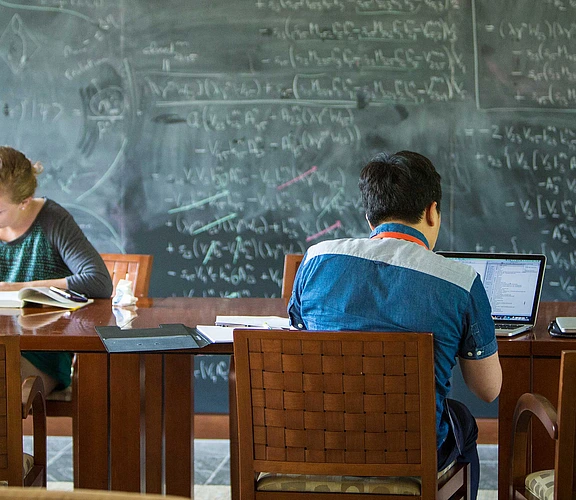 Scholars working at a table in front of a large chalkboard full of calculations.
