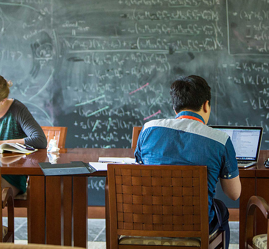 Scholars working at a table in front of a large chalkboard full of calculations.