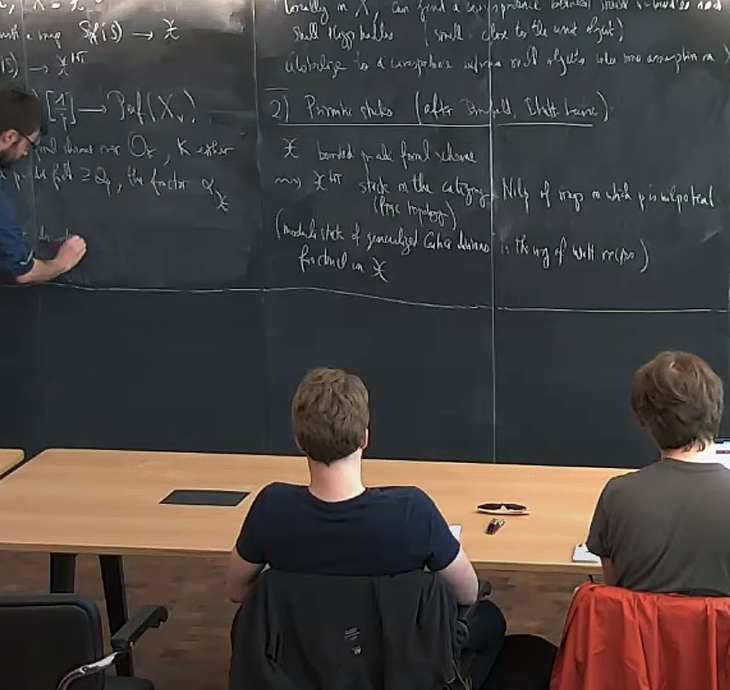 A screenshot of Arthur-Cesar Le Bras giving his talk on "A Stacky Perspective on P-adic Non-abelian Hodge Theory" at IAS.  He is writing on the chalkboard, and the view is from behind the first row of the audience.