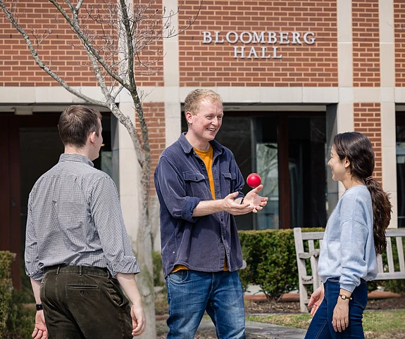 Three people standing outside Bloomberg Hall; Andy Mummery, at the center, juggles a red ball, smiling