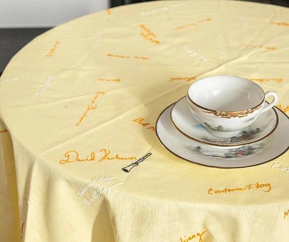 A white teacup with a gold rim sits on a matching saucer on a yellow tablecloth embroidered with various signatures in orange and white thread.