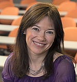 A headshot of Maria Chudnovsky. She is smiling, wearing a purple shirt, and standing in front of an auditorium.