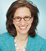 A professional headshot of Margaret Holen.  She is smiling, wearing glasses and a turquoise blazer, and sitting in front of a pale turquoise backdrop.