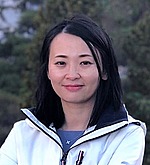 A headshot of Sui Tang.  She is wearing a white jacket over a blue shirt and standing outside.