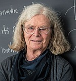 A headshot of Karen Uhlenbeck.  She is wearing a gray shirt, black scarf, and standing in front of a chalkboard.