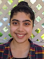 A headshot of Neha Wadia. She is smiling, wearing a plaid flannel shirt, and standing in front of white lattice.