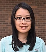 Headshot of Fan Wei.  She is smiling, wearing glasses and a light blue button-down, and standing in front of a brick wall.