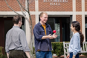 Three people standing outside Bloomberg Hall; Andy Mummery, at the center, juggles a red ball, smiling