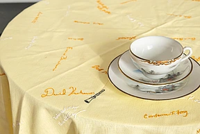 A white teacup with a gold rim sits on a matching saucer on a yellow tablecloth embroidered with various signatures in orange and white thread.