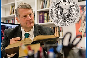 A man in a suit reads a large book in an office lined with bookshelves. An emblem for the American Philosophical Society overlays the image.