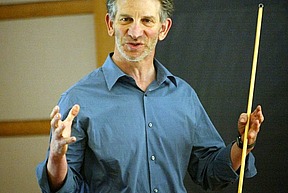 Peter Sarnak is standing in front of a chalkboard and lecturing to an unseen audience.  He is wearing a blue, button-down shirt and holding a pointer in his left hand.