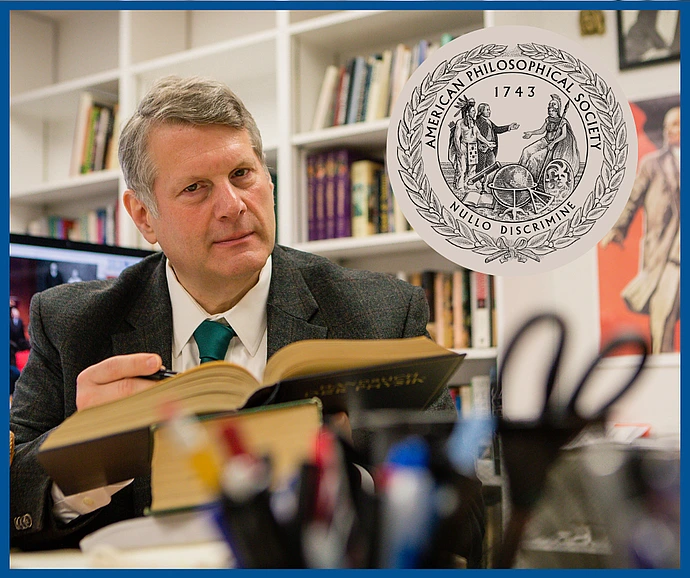A man in a suit reads a large book in an office lined with bookshelves. An emblem for the American Philosophical Society overlays the image.