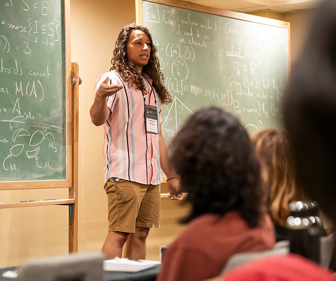 A lecturer discusses mathematical research in front of two green chalkboards