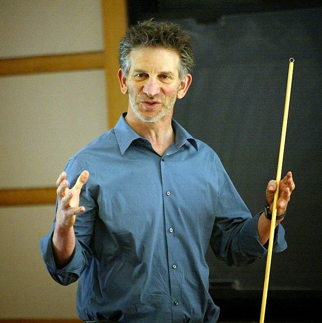 Peter Sarnak is standing in front of a chalkboard and lecturing to an unseen audience.  He is wearing a blue, button-down shirt and holding a pointer in his left hand.